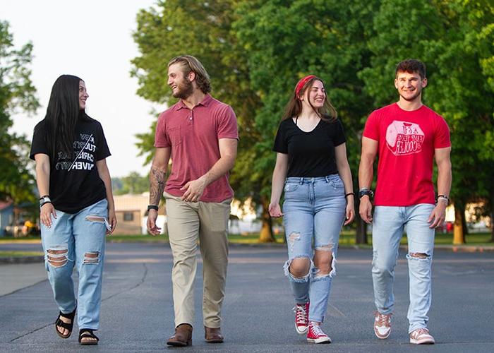 Four young adults walk and talk together outdoors on a paved path with greenery and sunlight in the background.