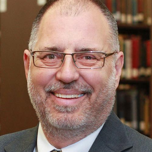 A smiling man with glasses, a beard, and wearing a suit is pictured in a library setting.