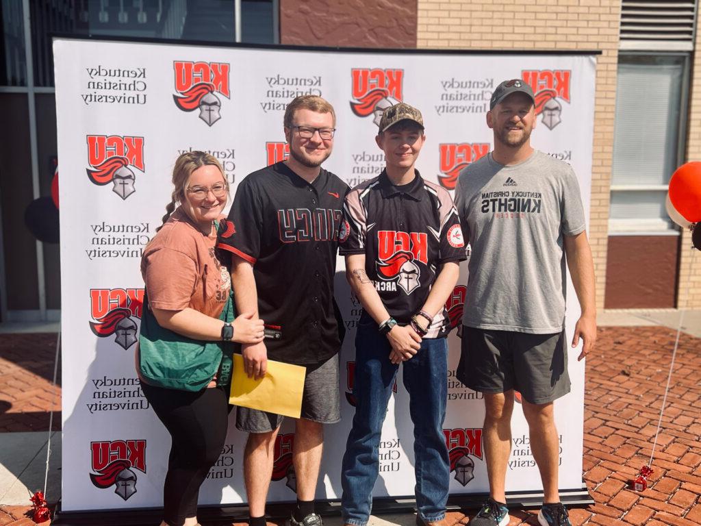 Four people stand smiling in front of a Kentucky Christian University backdrop outdoors, wearing university-themed shirts and casual clothing.